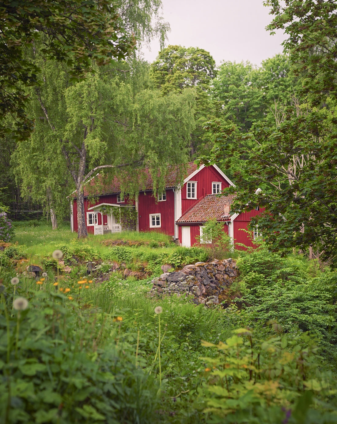 A red house nestled among green trees and wildflowers.